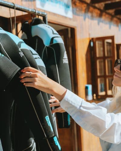 A woman reaching for a black wetsuit hanging on a rack inside a dive shop.