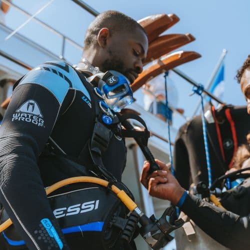 A diver in a wetsuit receiving assistance putting on their scuba gear from a crew member on a boat.