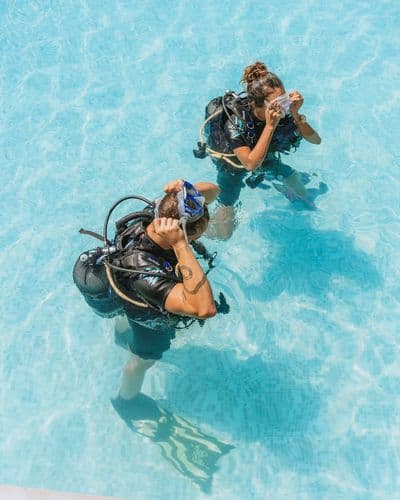 Two divers in a pool, wearing full gear, practicing mask clearing techniques underwater.